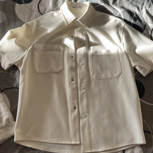 Vit kortärmad skjorta från Zara - Säljer en stilren vit kortärmad skjorta från Zara i storlek M. Skjortan har två stora bröstfickor och knappar framtill. Perfekt för sommardagar eller som en lätt overshirt. Materialet känns mjukt och bekvämt.