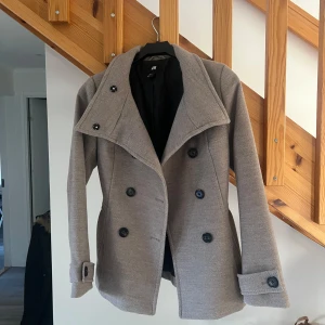 Grå/beige kappa  - Säljer en stilren kort kappa från H&M i mycket bra skick. Den har en dubbelknäppt design med stora svarta knappar och en klassisk krage. Perfekt för höst och vår! Passar till både vardag och lite finare tillfällen💕☁️ 