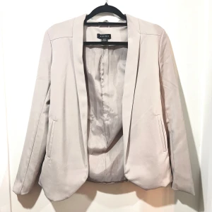 Grå beige kavaj - Snygg gråbeige kavaj med gulddetaljer baktill. Den har en stilren design med långa ärmar och en normal passform. Perfekt för både vardag och fest!