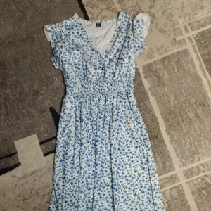 Blommig sommarklänning - Söt blommig klänning i vitt och blått med volangdetaljer. Perfekt för sommardagar och midsommarfirande. Klänningen har en omlott-design och är i mycket bra skick. Den är ärmlös och har en smickrande passform.