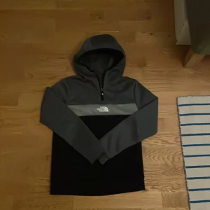 Grå och svart hoodie från The North Face - Säljer en snygg och bekväm grå och svart hoodie från The North Face. Perfekt för kyliga dagar med sin mjuka insida och praktiska huva. Den har en halv dragkedja framtill och är i mycket bra skick. Passar perfekt för både vardag och utomhusaktiviteter. Köpt på JD sports.