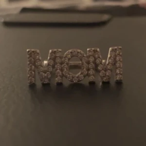 Ring med texten 'MOM' i strass - Snygg ring med texten 'MOM' dekorerad med glittrande strass. Perfekt som present till mamma eller för att visa uppskattning. Ringen har en öppen design vilket gör den justerbar i storlek. Passar till både vardag och festliga tillfällen.