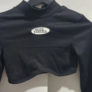 Svart ribbad croppad tröja från No Fear x H&M - Säljer en svart ribbad croppad tröja från No Fear x H&M. Tröjan har en cool logga framtill och långa ärmar med tumhål. Perfekt för en edgy look! 🖤