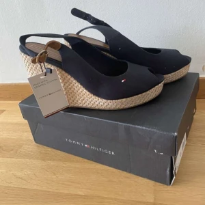 Svarta kilklackar från Tommy Hilfiger - Säljer ett par snygga svarta kilklackar från Tommy Hilfiger. Skorna är tillverkade av ekologisk bomull och har en flätad kilklack i naturfärgat material. De har en öppen tå och en rem runt hälen med ett litet spänne. Perfekta för sommaren och kan lätt matchas med både klänningar och jeans.