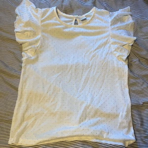 Vit topp med prickar från Lindex - Säljer en söt vit topp från Lindex med små prickar över hela tyget. Toppen har korta ärmar med volangdetaljer och en liten knappöppning i nacken. Perfekt för en somrig look! 🌸