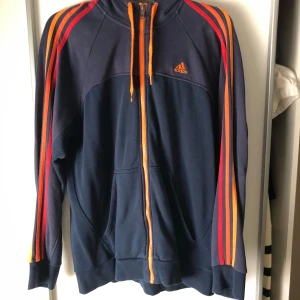 Blå Adidas jacka med orange detaljer - Säljer en blå Adidas hoodie med orange dragkedja och detaljer. Väl använd, bra skick. Storlek L men sitter som M.