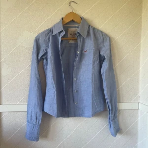 Blå skjorta från Hollister - Säljer en snygg blå skjorta från Hollister i storlek S. Skjortan är tillverkad av 98% bomull och 2% spandex, vilket gör den både bekväm och lite stretchig. Den har långa ärmar och knappar längs framsidan. Perfekt för både vardag och fest!