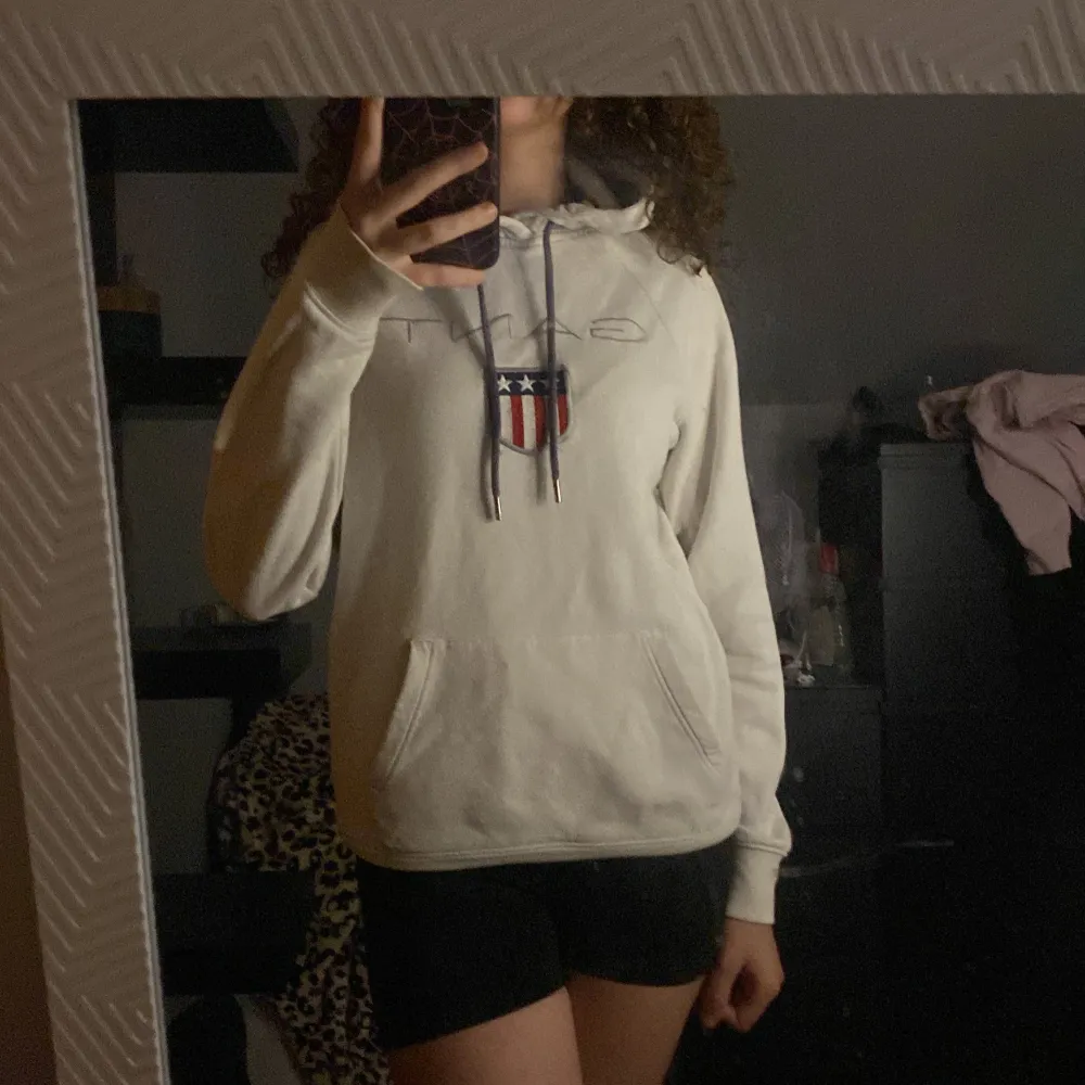 Säljer en vit hoodie från GANT i storlek XS. Hoodien har en stor ficka på magen och ett snyggt tryck med GANT-loggan Den har långa ärmar och en justerbar huva med snören. den är lite sliten på armbågen . Tröjor & Koftor.