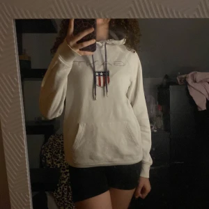 Vit hoodie från GANT - Säljer en vit hoodie från GANT i storlek XS. Hoodien har en stor ficka på magen och ett snyggt tryck med GANT-loggan Den har långa ärmar och en justerbar huva med snören. den är lite sliten på armbågen 