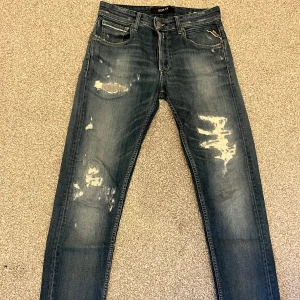 Blå slitna jeans från Replay - Säljer ett par snygga blå jeans från Replay med slitna detaljer på framsidan, modellen heter Grover.. De har en klassisk femficksdesign och en knappgylf. Perfekta för en avslappnad stil och passar till både vardag och fest.