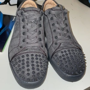 Louboutin buda!  - Säljer ett par svarta sneakers från Louboutin i mycket bra skick. Skorna har en cool design med nitar på tån och den klassiska röda sulan. Perfekta för att ge din outfit en edgy touch. De är i storlek herr 42-44 och har snörning för en bra passform.