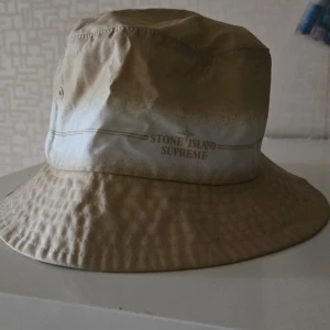 Beige bucket hat från Stone Island x Supreme - Snygg beige bucket hat från Stone Island x Supreme. Perfekt för sommardagar med sin lätta och bekväma passform. Hatten har en diskret logga framtill och är i mycket bra skick. Ett måste för streetwear-entusiaster!