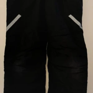 Svarta överdragsbyxor från Woxo - Säljer ett par svarta överdragsbyxor från Woxo, designade av KappAhl. De har praktiska dragkedjor och reflexdetaljer som gör dem perfekta för utomhusaktiviteter. Snölås och justerbar kardborre längst ner. Justerbar kardborre på utsidan i midjan. Två sidfickor med blixtlås. Stängs med blixtlås och knappar i midjan.   Reflexer på benen. Finns hällor för hängslen i midjan som tyvärr inte medföljer.  Något slitna på jämnan och början till ett hål på höger knä (se sista bild). 