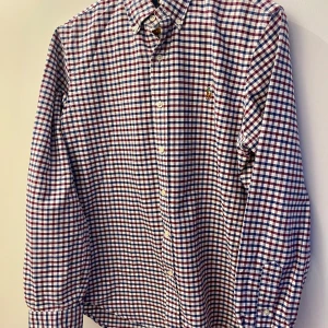 Klassisk rutig Ralph Lauren-skjorta – stilren & tidlös - Stilig och klassisk rutig skjorta från Ralph Lauren i vitt, blått och rött.                                                                    Med långa ärmar och knäppning framtill är den enkel att bära både till vardags och festliga tillfällen.                En tidlös skjorta i toppskick!