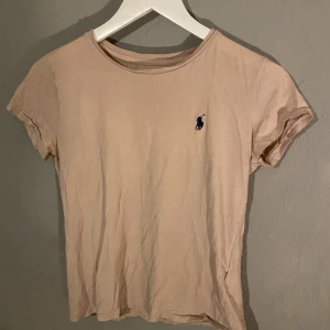 Beige/rosa t-shirt från Ralph Lauren - Säljer en snygg beige/ljusrosa t-shirt från Ralph Lauren med deras klassiska logga broderad i blått på bröstet. T-shirten är kortärmad, perfekt för en avslappnad stil. Passar bra till både jeans och shorts! (PM OM DU VILL HA FLER BILDER)