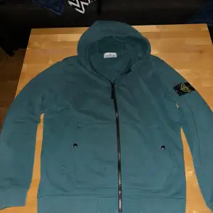 Säljer en snygg grön hoodie från Stone Island. Den har en dragkedja framtill och en klassisk huva. Perfekt för höst och vår, med långa ärmar och en bekväm passform. Märkets ikoniska patch finns på ärmen. Perfekt för en casual look!