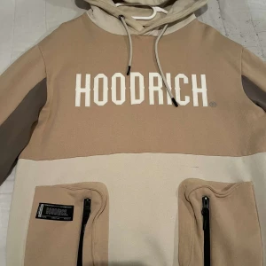 Beige och brun hoodie från Hoodrich - Säljer en snygg beige och brun hoodie från Hoodrich i storlek S. Den har en stor logga på bröstet och praktiska fickor med dragkedja. Perfekt för höst och vår, och den är i mycket bra skick. Huvan har snören för justering. Skön och stilren för vardagsbruk!