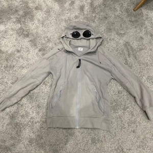 C.P. Company ziphoodie  - Säljer en unik beige huvtröja från C.P. Company med inbyggda glasögon i huvan. Tröjan har dragkedja framtill och två fickor med dragkedjor. Perfekt för höst och vår, och ger en cool look med sin speciella design. Passar både till vardags och lite mer avslappnade tillfällen.