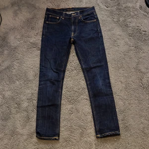 Mörkblå jeans från Nudie Jeans - Säljer ett par snygga mörkblå jeans från Nudie Jeans i mycket bra skick. De har en klassisk femficksdesign med orange sömmar och en läderpatch med logga baktill. Perfekta för både vardag och fest! Aldrig använda.