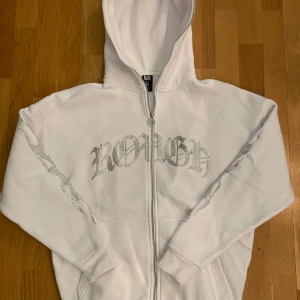 Vit hoodie från Black Squad - Säljer en vit hoodie från Black Squad i storlek XS. Den har en oversized passform och coolt tryck med texten 'ROUGH' i glittriga bokstäver på framsidan. Perfekt för en avslappnad stil med dragkedja och huva. Använd ett par gånger men i mycket bra skick. Perfekt för höst och vår!