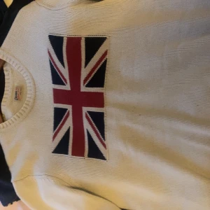 Vit tröja med UK logga på bröstet - Säljer en snygg vit tröja från Gant med ett stort Union Jack-mönster på framsidan. Tröjan är i bra skick och perfekt för både vardag och fest. Den har en klassisk passform och är tillverkad i ett mjukt material som känns skönt mot huden.