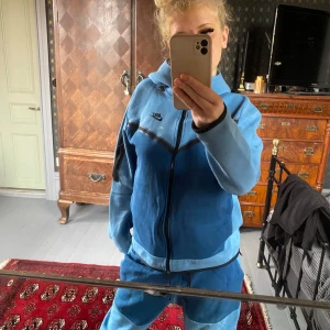 Blå träningsset från Nike - Säljer ett snyggt blått träningsset från Nike. Setet består av en hoodie och matchande byxor. Perfekt för träning eller en chill dag hemma. Materialet är mjukt och bekvämt, och det finns en dragkedja på hoodien. Byxorna har en elastisk midja med snörning för bästa passform. Fläckar på både jackan och byxorna. Väldigt speciell design o få har den här färgen. Storlek L