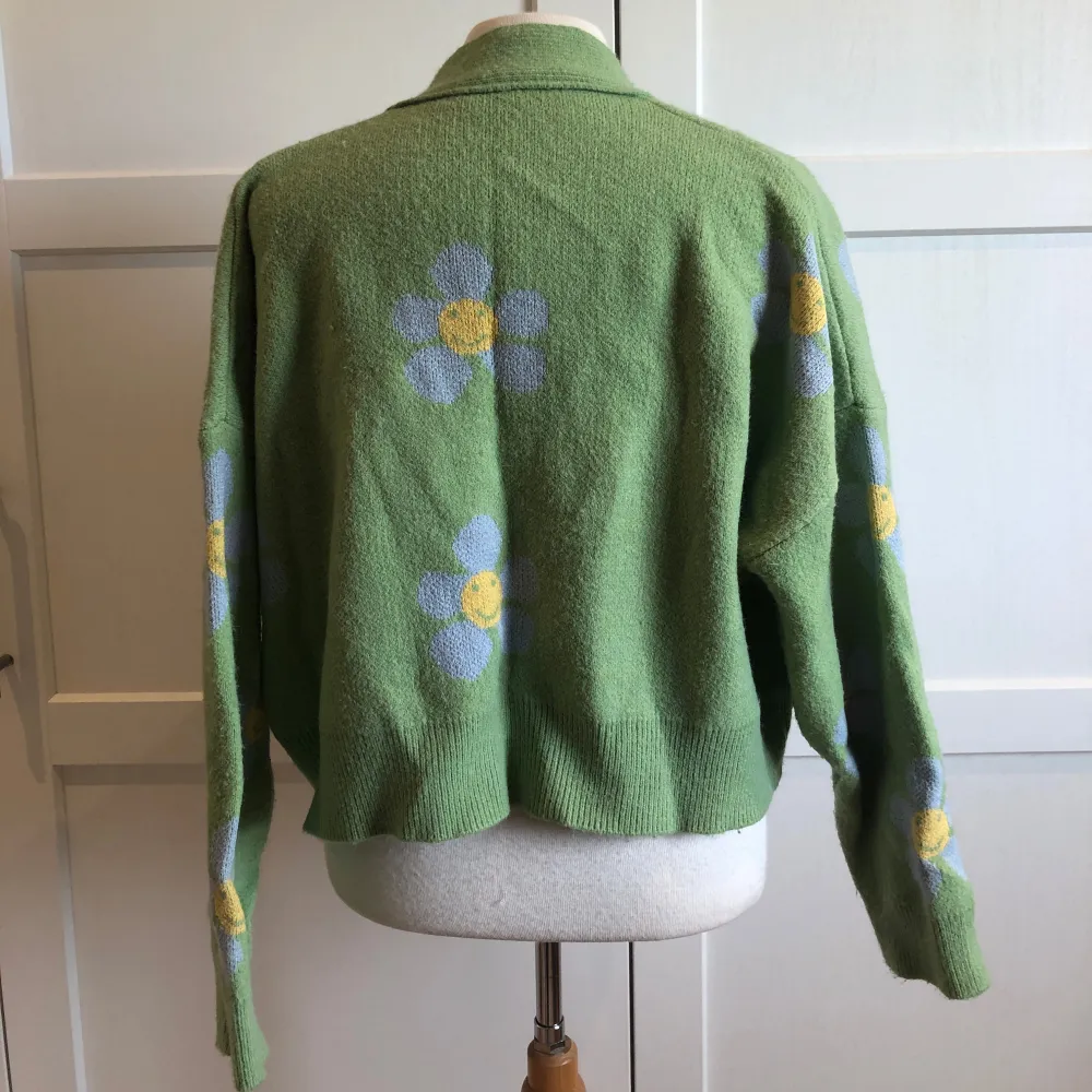 Superfin grön kofta med glada blommor i vitt och gult. Den går ner till höften och har stora vita knappar framtill. Perfekt året om, och ger en härlig retrovibe. Ärmarna är långa och koftan är i mycket bra skick. Mjuk och skön.. Neuletakit & Villapaidat.