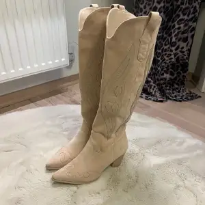 Säljer ett par snygga beige cowboy boots i mocka. De har en spetsig tå och en låg klack som ger en cool look. Perfekta för höst och vår, och de har fina broderade detaljer som gör dem unika. Passar perfekt till både jeans och klänning! Använda 1 gång