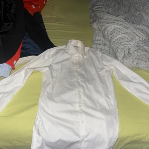 Vit skjorta och kavaj - Säljer en klassisk vit skjorta i bomull, perfekt för både formella och avslappnade tillfällen. Skjortan har långa ärmar och knappar framtill. Den är i bra skick och passar utmärkt till kostym eller jeans. Perfekt för vår och sommar! Köpt på nicco inför studenten 2022 använd endast en gång! Som ny köpt för 2200 kan självklart diskutera priset 