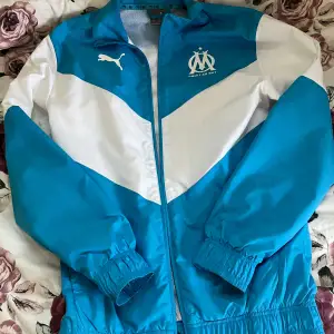 Säljer en snygg blå och vit jacka från Puma med Olympique de Marseille-logga. Jackan har dragkedja och långa ärmar, perfekt för sportiga tillfällen eller som en casual look. Den är i bra skick och redo för nya äventyr! 💙