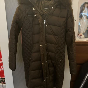 Brun dunjacka med pälskrage - Säljer en snygg brun dunjacka från Rock and Blue. Den har en mysig pälskrage. Perfekt för kalla vinterdagar med sin långa design och dragkedja framtill. Jackan har också praktiska fickor och är i mycket bra skick.