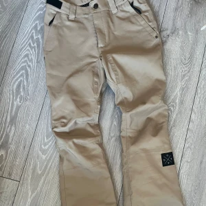 Beige täckbyxor från Dope - Säljer ett par snygga beige täckbyxor från Dope i storlek S. Byxorna har en smal passform och är perfekta för äventyr i snön. De är tillverkade i ett slitstarkt material och har en liten logga på benet.
