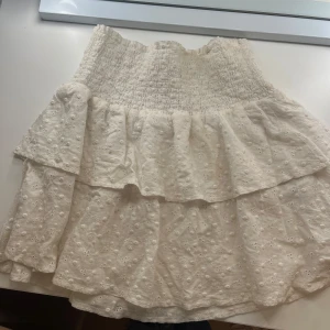 Vit kjol från Chelsea - Superfin vit kjol från Chelsea i storlek XS. Den har en smockad midja och volanger som ger en söt och romantisk look. Perfekt för sommardagar eller en mysig vårutflykt. Kjolen är i mycket bra skick och har ett diskret blommönster som ger extra charm. Passar perfekt med en stickad tröja eller en enkel topp!