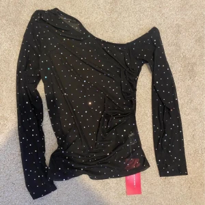Svart oneshoulder topp med glitter - Snygg svart oneshoulder topp med glittriga detaljer. Perfekt för festliga tillfällen! Den har långa ärmar och en tight passform som framhäver figuren. Ny med tag kvar.