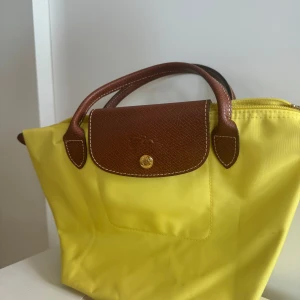 Gul handväska från Longchamp - Säljer en snygg mindre gul handväska från Longchamp i mycket bra skick. Väskan har bruna skinnhandtag och en matchande flik med tryckknapp. En klassisk och stilren väska som passar till det mesta! Nästan aldrig använd. 
