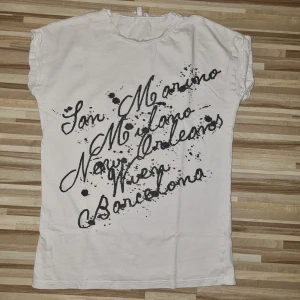 Vit t-shirt från Skill - Säljer en vit t-shirt från Skill i storlek 134/140. Den har ett coolt tryck med namn på olika städer som San Marino, Milano och Barcelona i svart. Den har 2 små hål fram i nedre kant och ett bak på vänster axel.