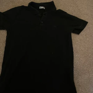 Svart pikétröja från Lacoste - Säljer en klassisk svart pikétröja från Lacoste i bra skick. Den har en stilren design med knappar framtill och den ikoniska krokodilloggan på bröstet. Perfekt för både vardag och lite mer uppklädda tillfällen. Passar till allt!
