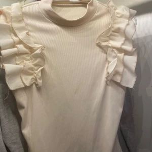 Beige ribbad topp med volangdetaljer - Säljer en superfin beige ribbad topp med volangdetaljer längs axlarna. Toppen har en hög hals och är ärmlös, vilket gör den perfekt för både vardag och fest. Den är i ett mjukt och stretchigt material som sitter bekvämt på kroppen.