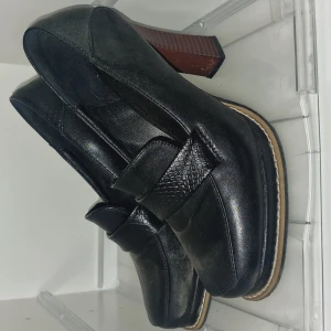 Svarta loafers med klack - Snygga svarta loafers i skinn med en robust klack. De har en elegant remdetalj över foten och en bekväm passform. Perfekta för både vardag och festliga tillfällen. Klacken ger en extra touch av stil och höjd.