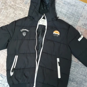 Svart pufferjacka från A. Hammond - Säljer en svart pufferjacka från A. Hammond i storlek M. Jackan har coola broderade detaljer och märken på bröstet och ärmen. Den är långärmad med dragkedja och har en skön huva. Perfekt för höst och vinter!