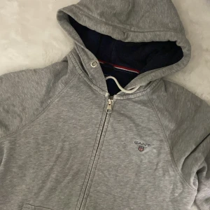 Grå hoodie från GANT - Säljer en snygg och bekväm grå hoodie från GANT. Den har en dragkedja framtill och en klassisk känguruficka. Perfekt för kyliga dagar eller mysiga hemmakvällar. Huvan är fodrad för extra värme och komfort. Passar till det mesta i garderoben!