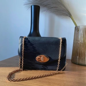 Svart axelväska med kedja - Snygg svart axelväska i skinn med guldfärgad kedja och ett elegant lås framtill. Perfekt för både vardag och fest. Väskan har en kompakt design som gör den lätt att bära med sig. Passar till alla säsonger och ger en stilren touch till din outfit.