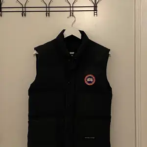 Säljer en svart dunväst från Canada Goose i mycket bra skick. Västen har en klassisk design med dragkedja och knappar framtill samt en broderad logotyp på bröstet. Perfekt för kyliga höst- och vinterdagar. Passar både till vardags och mer uppklädda tillfällen.