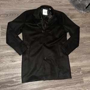 Svart rock från Produkt - Snygg svart rock från Produkt i klassisk design. Den är är storlek M.Rocken har en stilren look med knappar framtill och är perfekt för kyligare dagar. Den är långärmad och har en elegant passform som passar både till vardags och mer formella tillfällen.