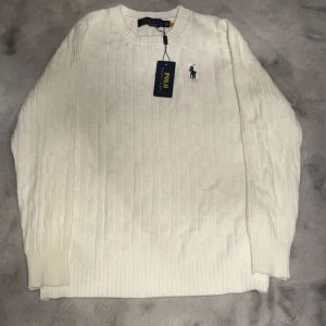 Vit tröja från Ralph Lauren - Säljer en stilren vit tröja från Ralph Lauren med kabelstickat mönster och den klassiska loggan broderad på bröstet. Tröjan har långa ärmar och en rund halsringning. Perfekt för en elegant och avslappnad look. Storlek s