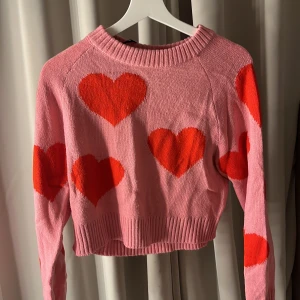 Rosa stickad tröja med hjärtan - Säljer en söt rosa stickad tröja med stora röda hjärtan. Perfekt till alla hjärtans dag som är runt hörnet!Tröjan är croppad och har långa ärmar med ribbade kanter. Perfekt för en mysig och lekfull stil. 💖