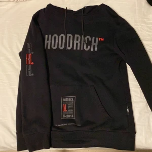Svart hoodie från Hoodrich - Säljer en svart hoodie från Hoodrich med stor logga på bröstet och ryggen. Hoodien har en känguruficka framtill och justerbar huva med dragsko. Perfekt för en avslappnad stil.