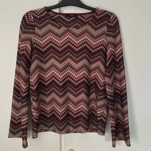 Mönstrad långärmad topp från Lindex - Säljer en snygg långärmad topp med vida armar från Lindex med ett zigzag-mönster i vinrött, rosa och beige. Perfekt för att liva upp garderoben med lite färg och mönster. Passar bra till både jeans och kjol. Hör av dig vid intresse!