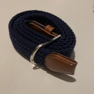  flätat bälte  - Snyggt blått flätat bälte med bruna skinn-detaljer och en silverfärgad spänne. Perfekt för att ge en stilren touch till din outfit. Bältet har en flexibel design tack vare det flätade materialet.