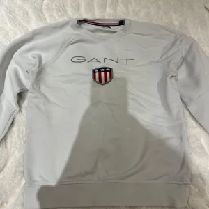 Vit sweatshirt från GANT - Säljer en stilren vit sweatshirt från GANT med ett broderat emblem i rött, vitt och blått på bröstet. Tröjan har rund hals och långa ärmar, perfekt för en avslappnad stil. Passar bra till både jeans och chinos.